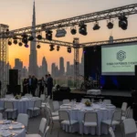 Outdoor corporate event setup in Dubai with stage, lighting, branded screen, and city skyline at sunset.